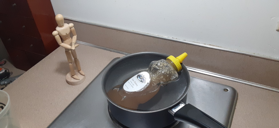 a bottle of honey in a saucepan of warm water. it's one of those bear-shaped bottles and its face is sticking out of the water, so it looks like it's enjoying a bath.  next to it is a drawing figurine, posed to look like it's waiting for the honey